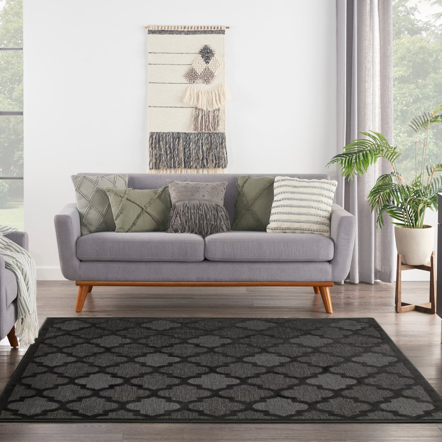 Gray mid-century sofa with light wood legs, green cushions, wall tapestry, potted plant, and a black geometric rug.
