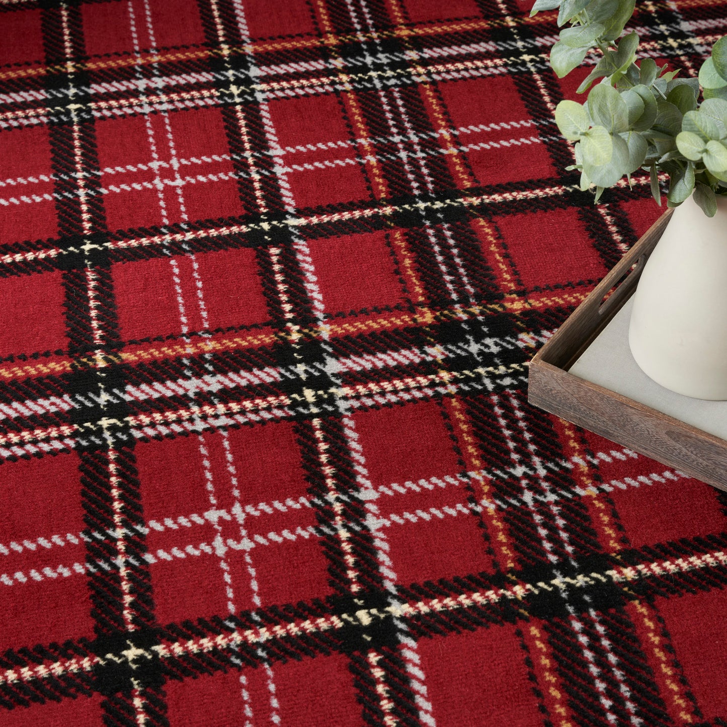 Red plaid rug with black, white and gold lines; wooden tray with a white vase and eucalyptus sits in the corner.