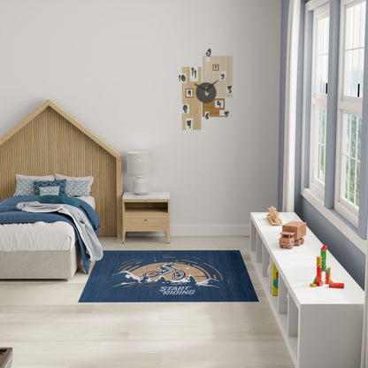 Kids' bedroom with a light wood house bed, navy Start Riding rug, white nightstand, blocks, wall clock and windows.