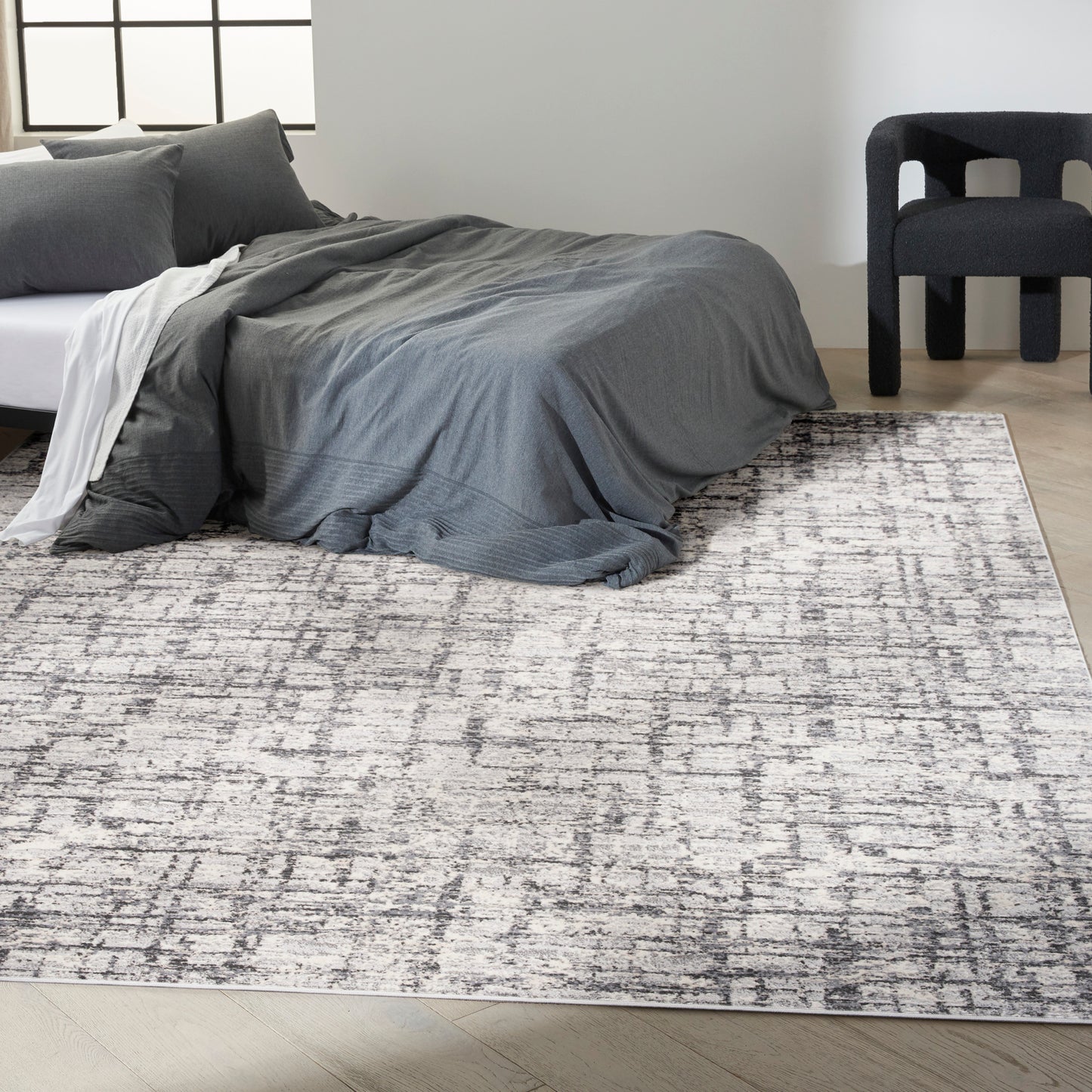 Gray duvet and pillows on a low bed with a white throw, atop a light gray abstract rug; dark curved chair nearby.
