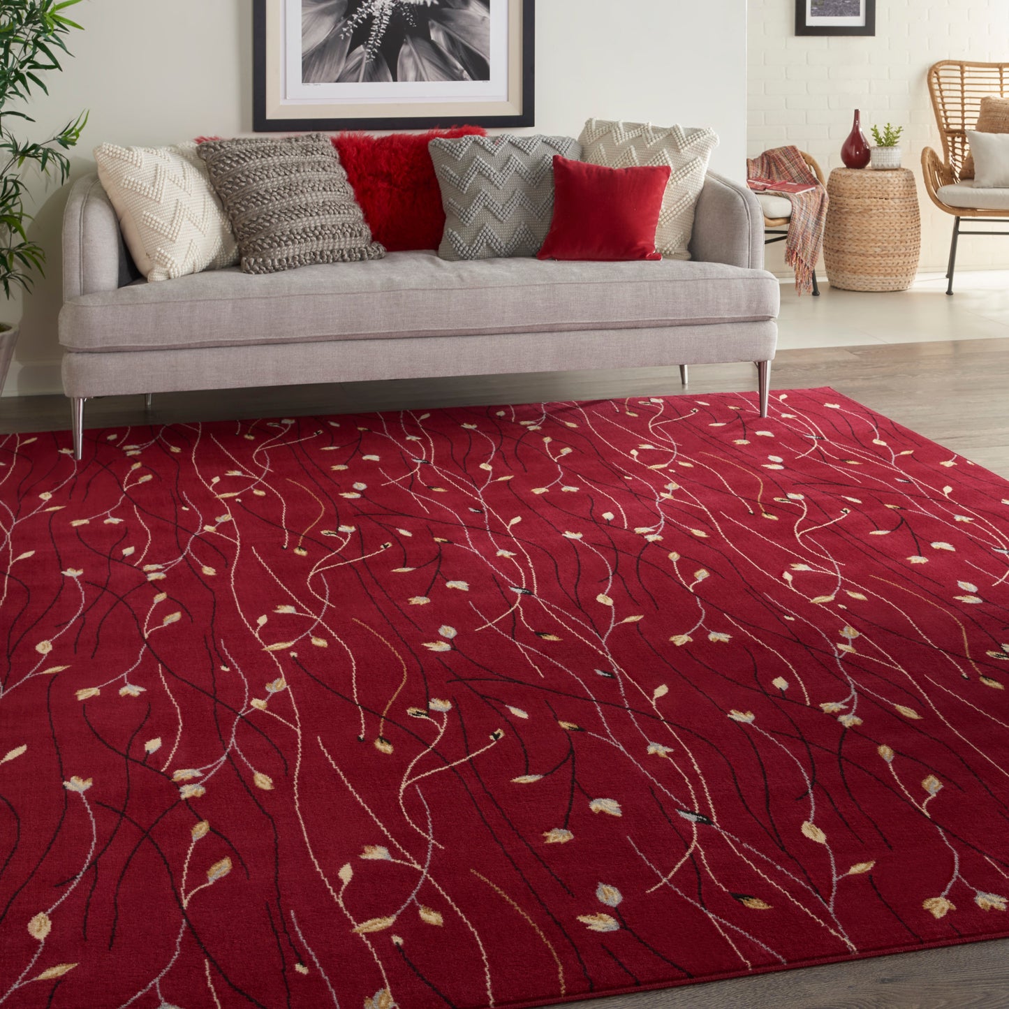 Red area rug with white vine pattern and small beige leaves under a light gray sofa with beige and red cushions.