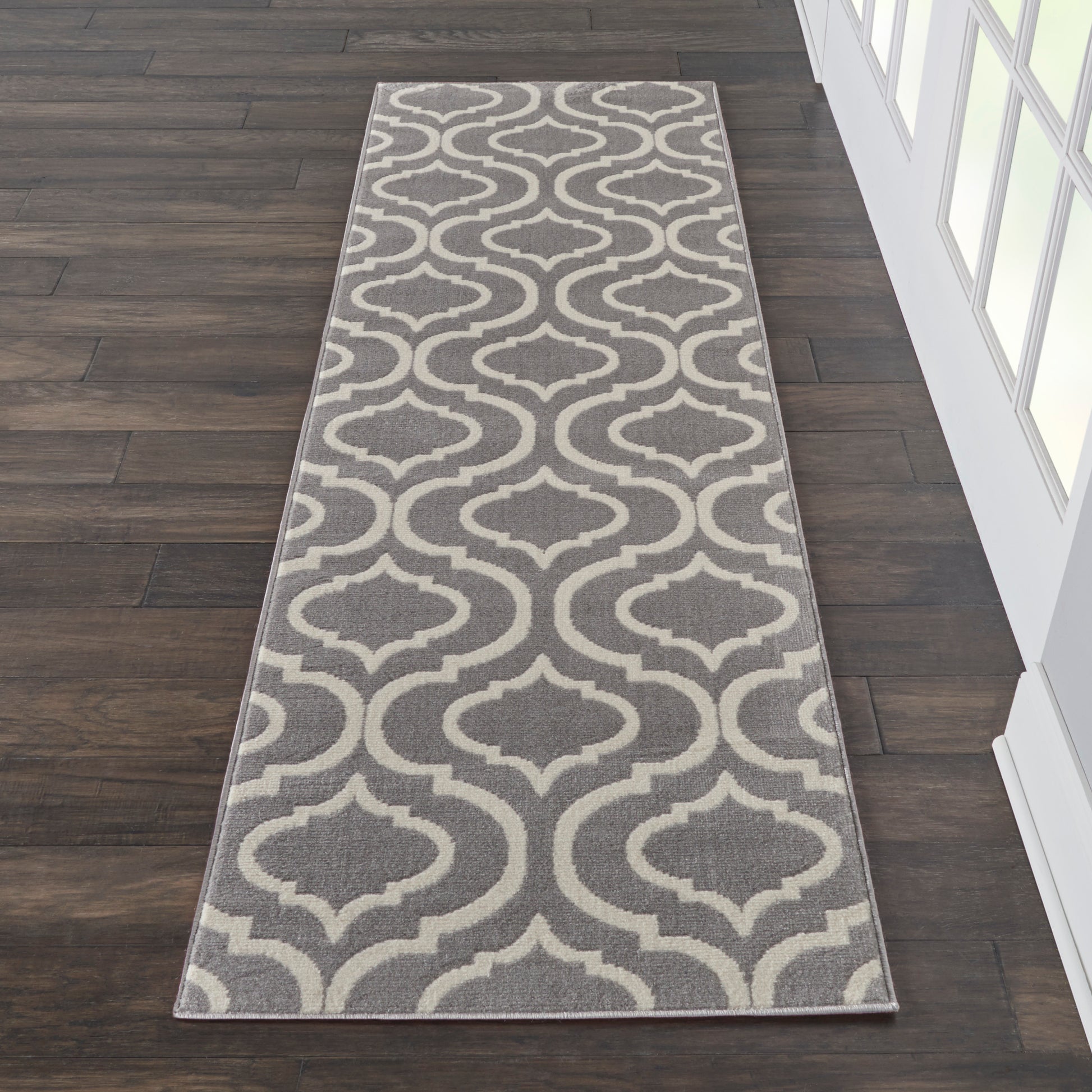 Grey & beige trellis-patterned 7' long runner rug on dark wood flooring beside a glass-paneled wall.