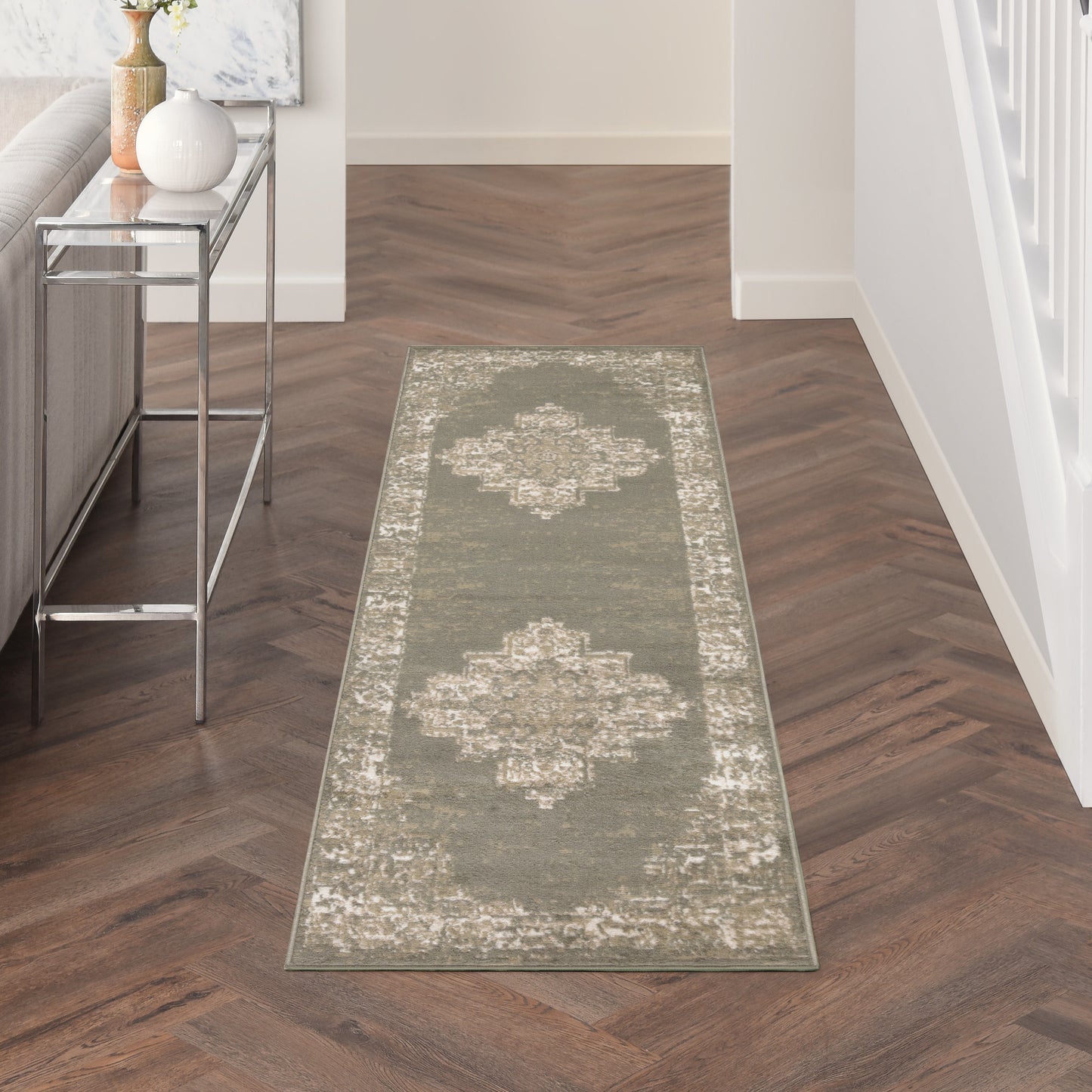 Olive-gray distressed runner rug with pale border in a hallway; dark herringbone floor and a chrome-glass console table nearby.