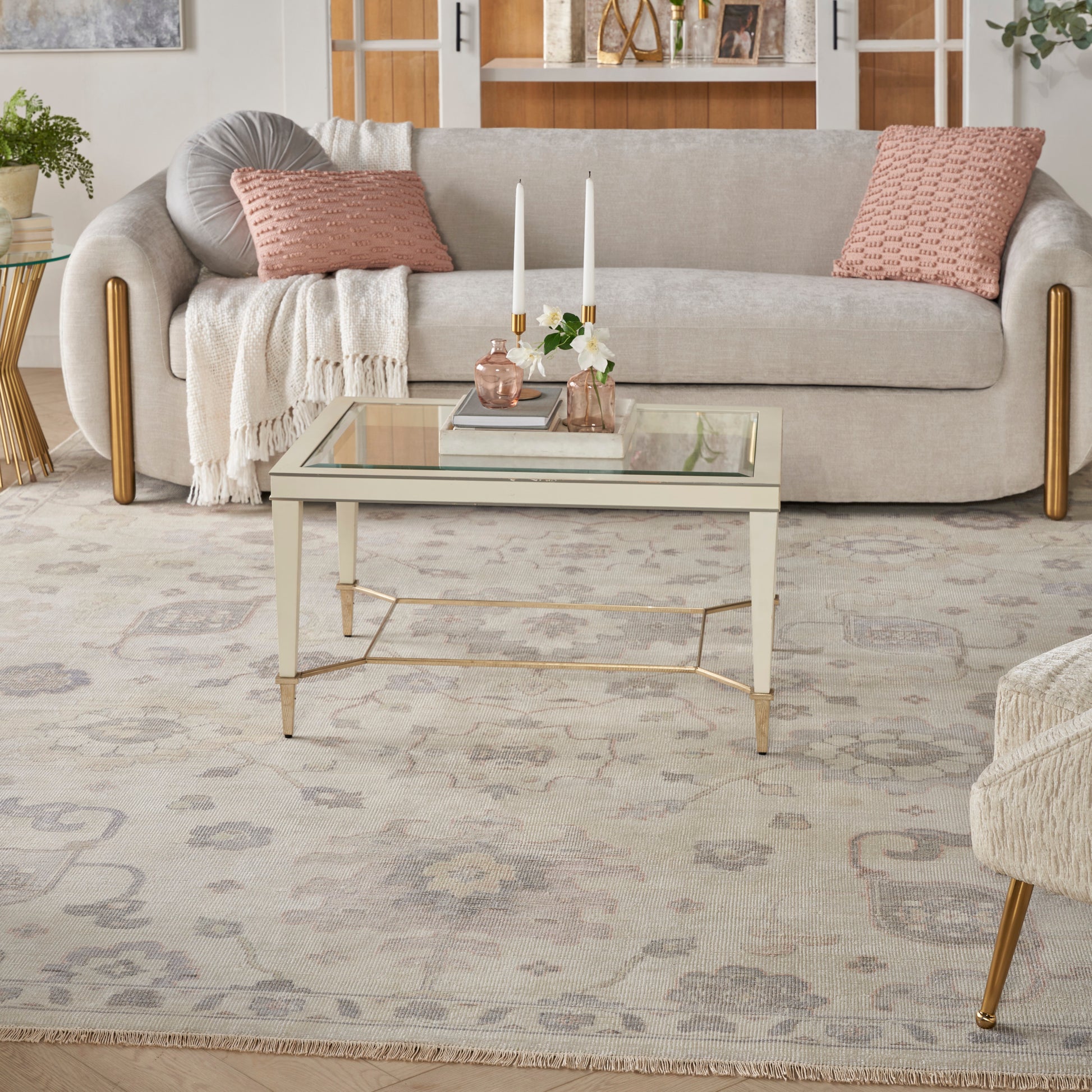 Beige fabric sofa with pink cushions and cream throw, glass coffee table with gold frame, soft patterned rug.