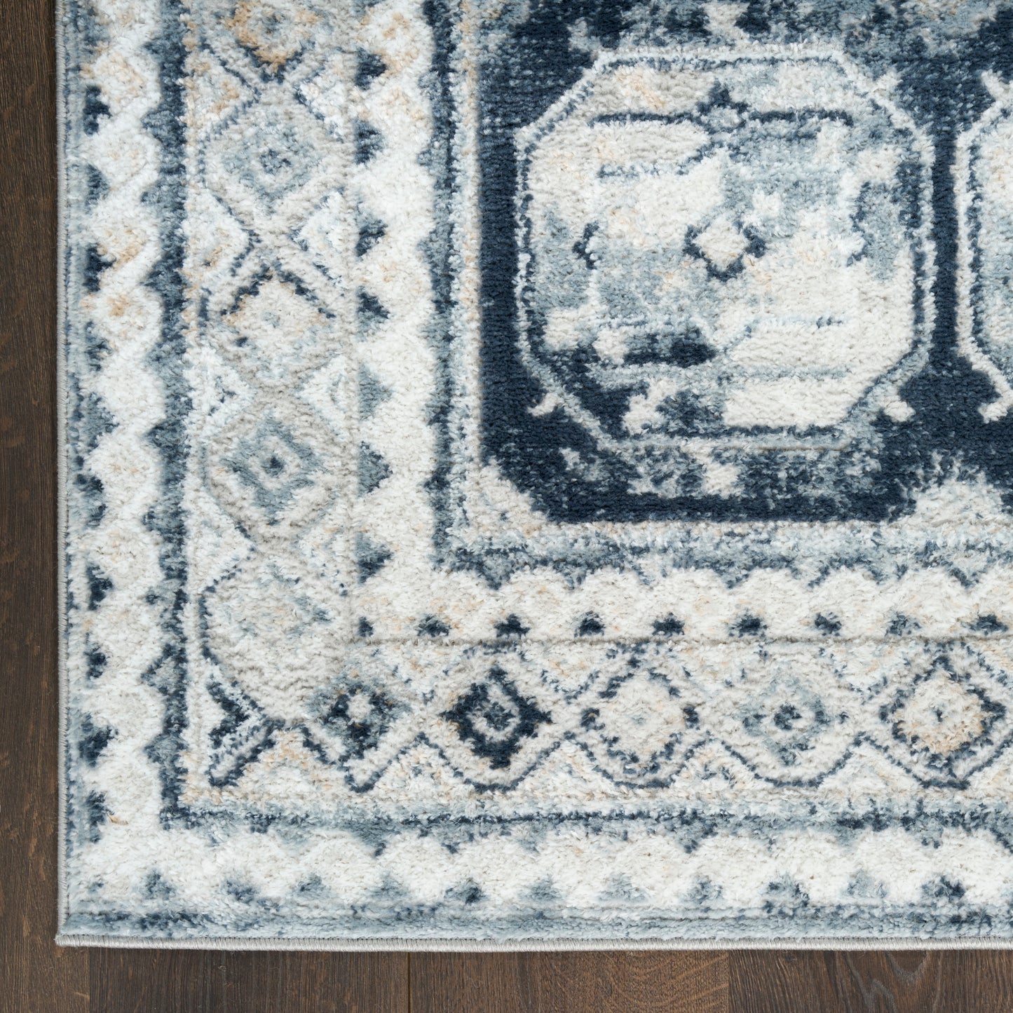 Blue, ivory and gray geometric area rug with diamond motifs and ornate border on dark wood floor.