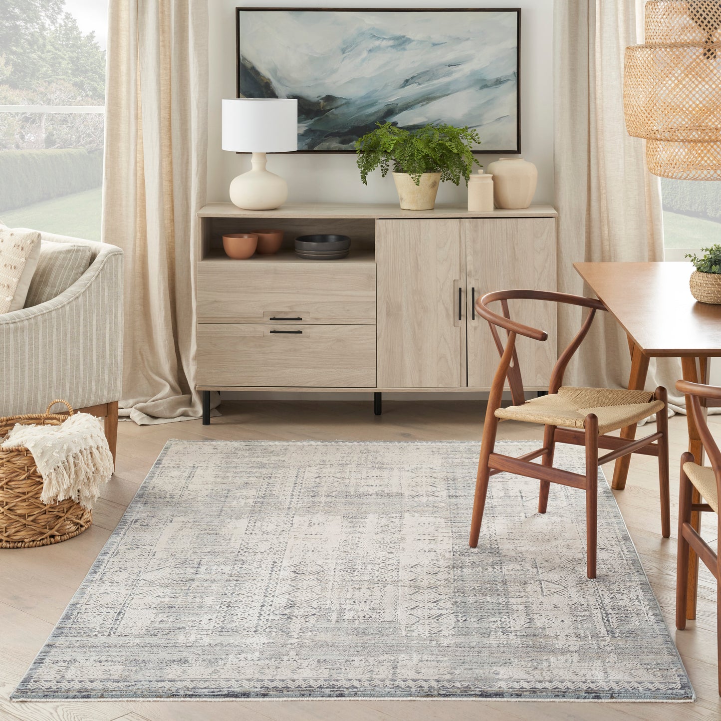 Light wood sideboard with plant, lamp, abstract wall art, blue-gray rug and wooden dining chairs in a bright living room.