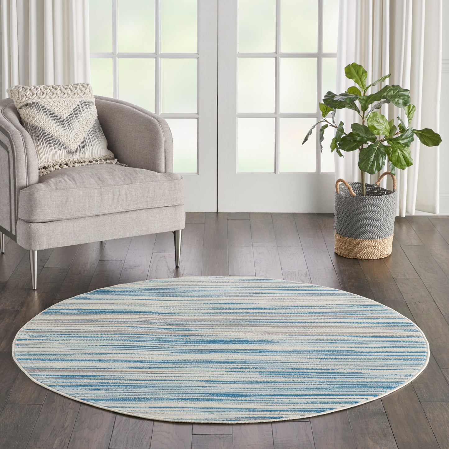 Round blue striped area rug (5' round) on dark wood floor, with a gray upholstered armchair and a potted plant in a woven basket by French doors.