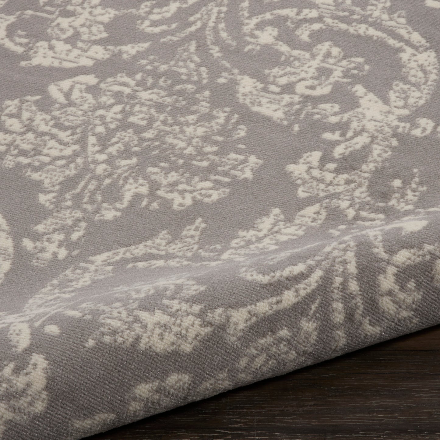 Taupe damask-patterned rug with ivory floral motifs, folded edge resting on a dark wooden floor.
