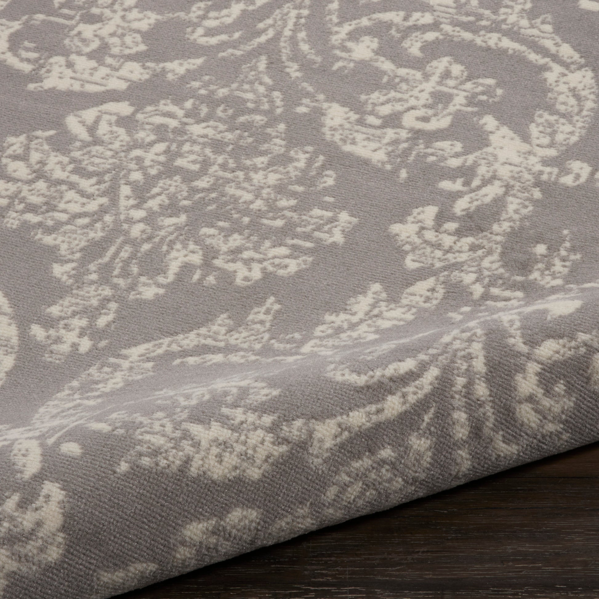 Taupe damask-patterned rug with ivory floral motifs, folded edge resting on a dark wooden floor.