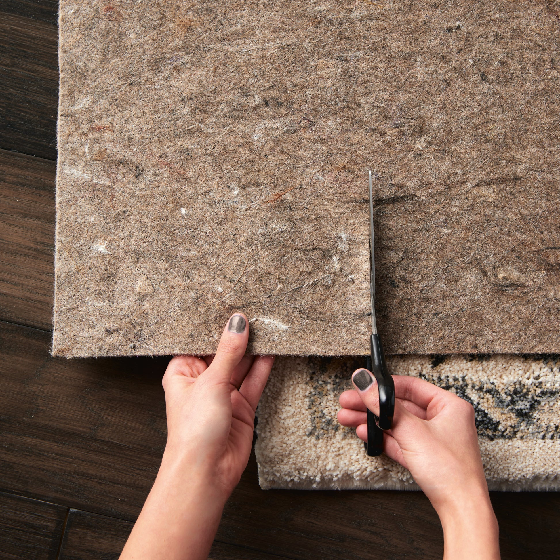 Beige textured area rug being trimmed with scissors by hands on dark wooden floor.