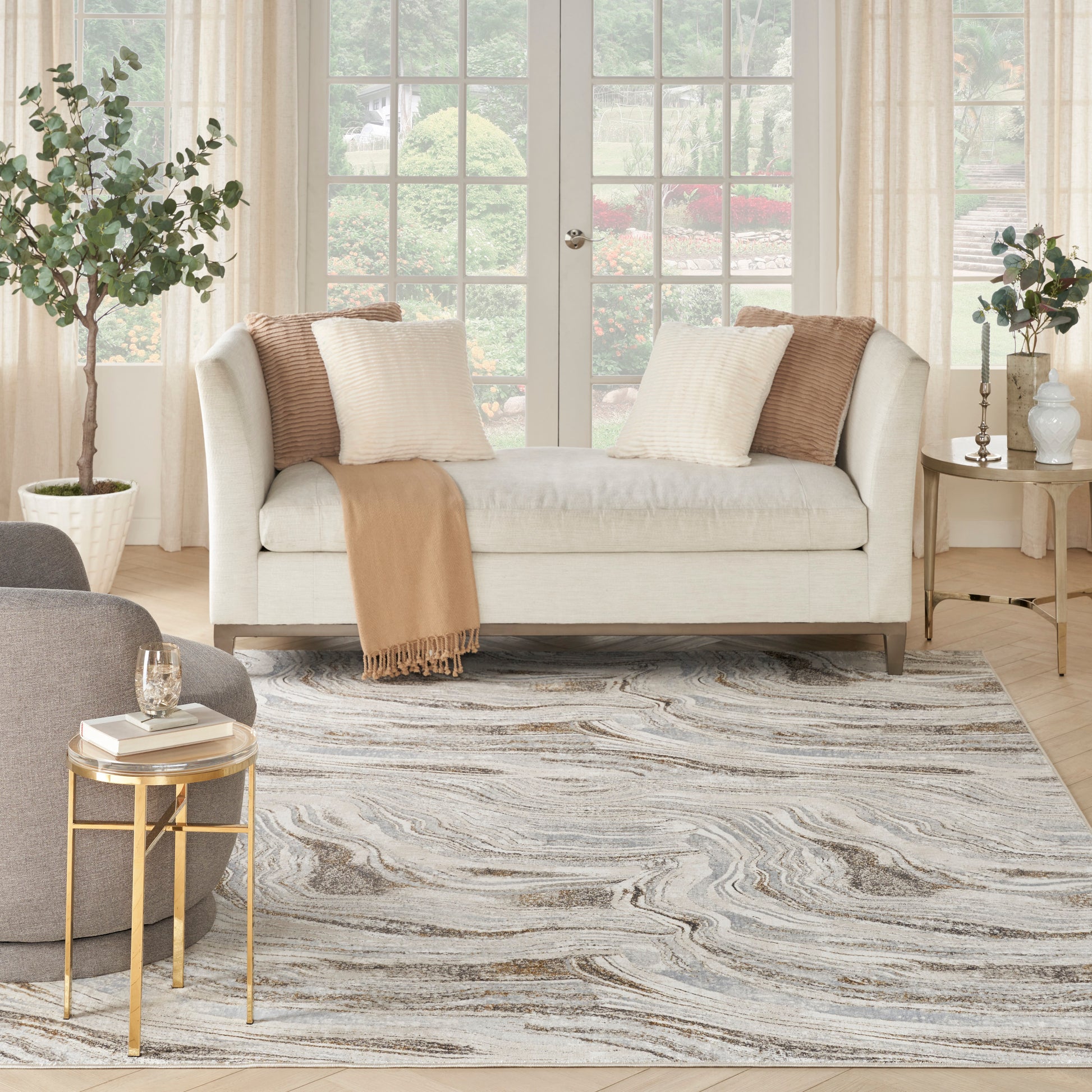 Swirled gray-blue area rug (8' x 10') in a living room with a beige sofa, cream & tan cushions, camel throw, gold side tables, potted plants and windows.