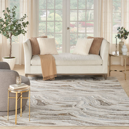 Swirled gray-blue area rug (8' x 10') in a living room with a beige sofa, cream & tan cushions, camel throw, gold side tables, potted plants and windows.