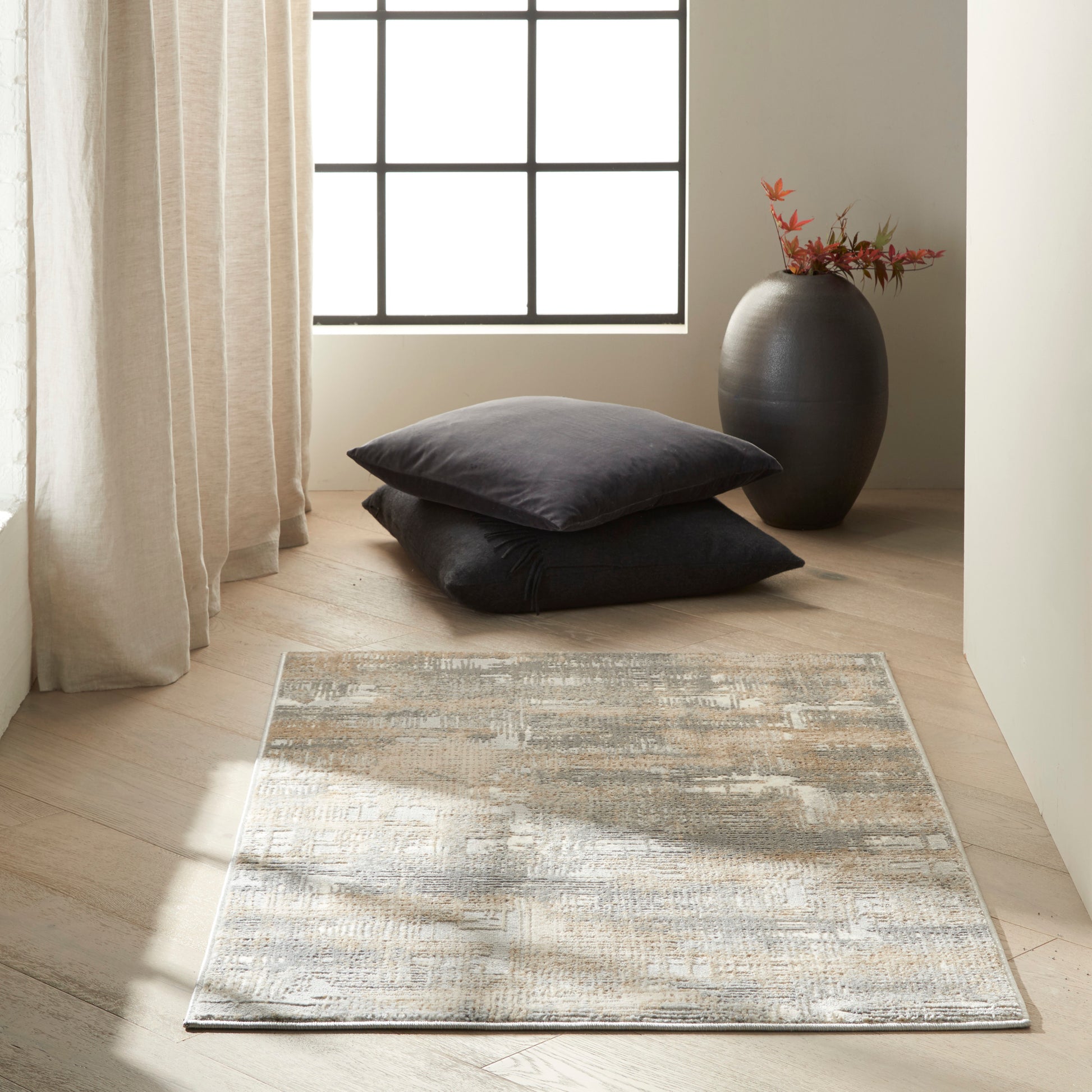 Neutral corner with grid window, beige curtains, light wood floor, gray abstract rug, stacked charcoal cushions, and a dark vase with red leaves.