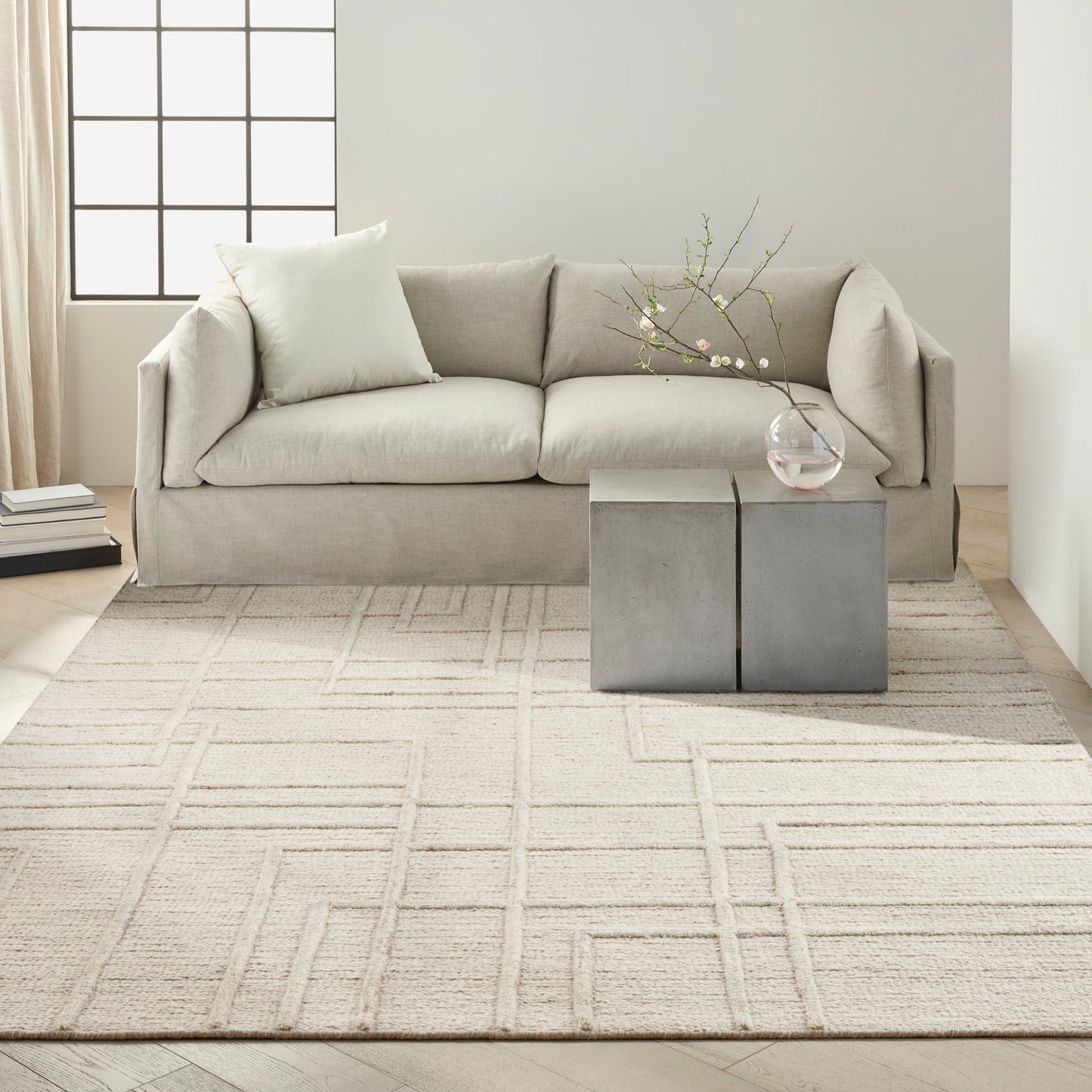 Beige fabric sofa with two cushions in a modern living room, gray cube side tables, glass vase with branches & a light grid-pattern rug.