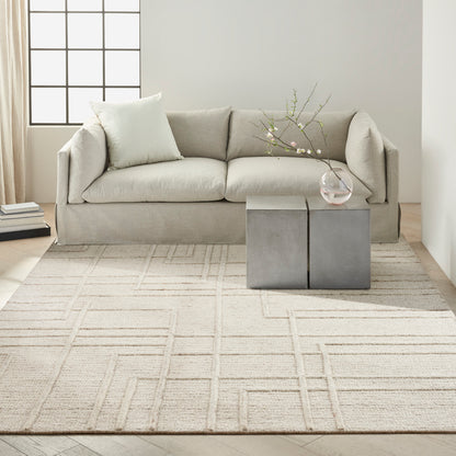 Beige fabric sofa with two cushions in a modern living room, gray cube side tables, glass vase with branches & a light grid-pattern rug.