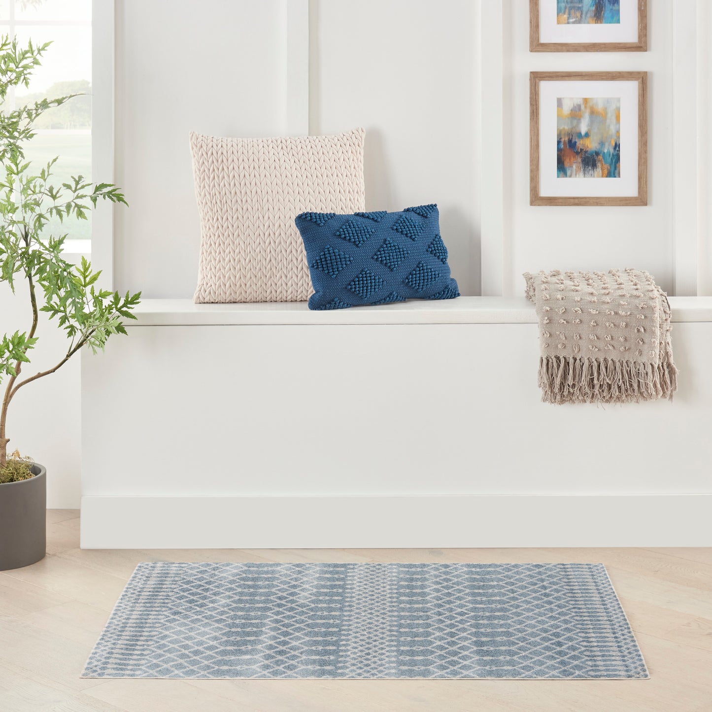 Bright living with white bench, beige knit cushion, blue geometric cushion, beige fringe throw, blue rug, plant and art.