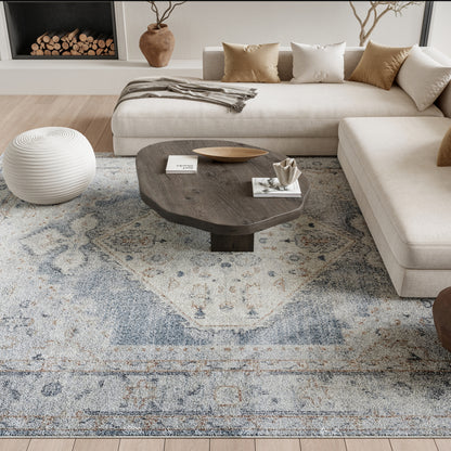 Beige sectional with dark wood coffee table on blue patterned rug; round white woven pouf and tan cushions.