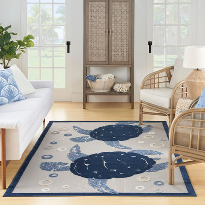 Rectangular light-blue woven rug with navy sea turtles; coastal living room with white sofa and wicker chairs.
