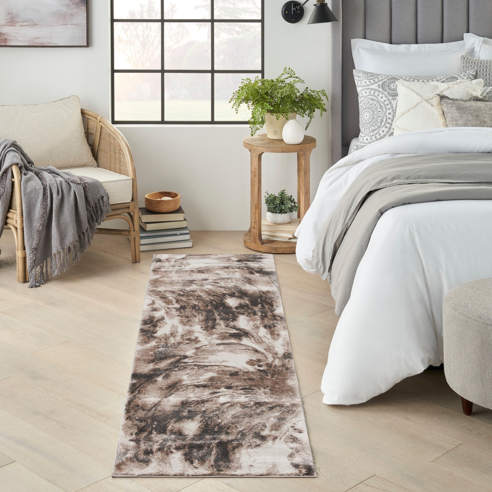 Long brown-beige abstract runner rug on light wood floor beside a white bed, wicker chair, and potted plant.