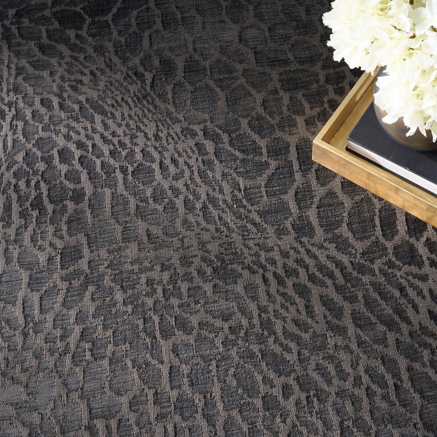 Dark gray animal-pattern textured rug with a light wood tray holding white flowers and books.