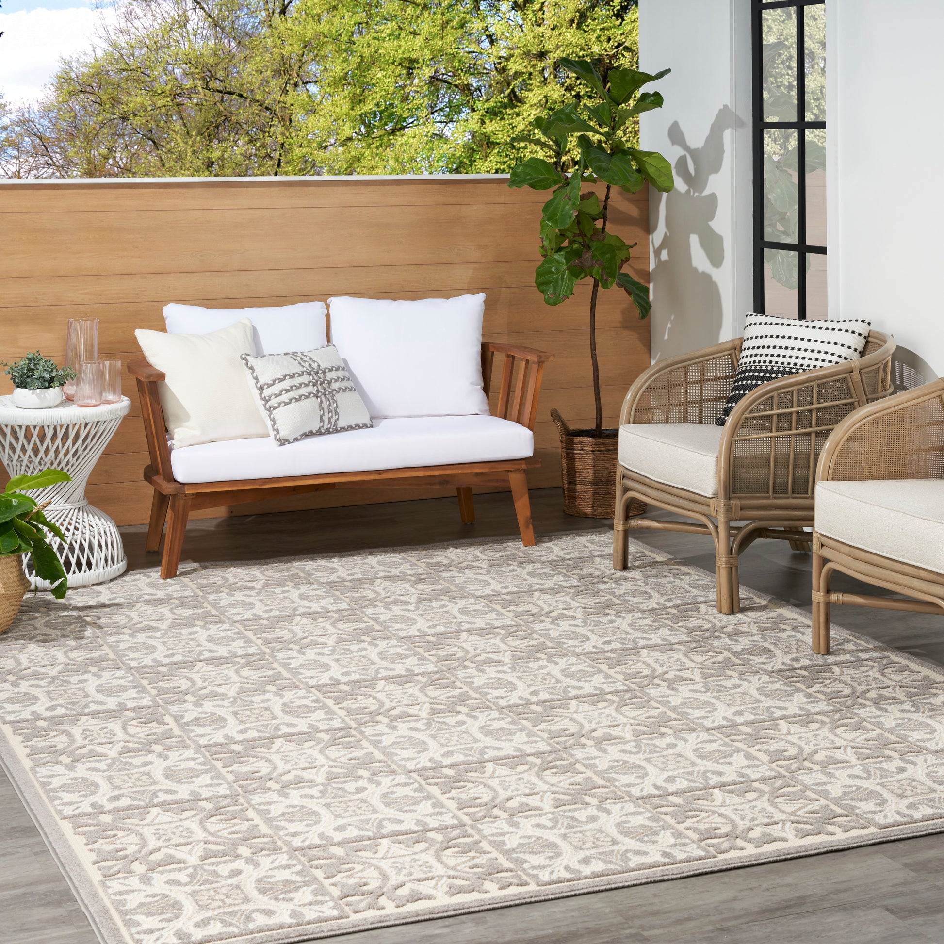 Teak outdoor sofa with white cushions, wicker armchairs, and a beige patterned outdoor rug on a sunny patio.