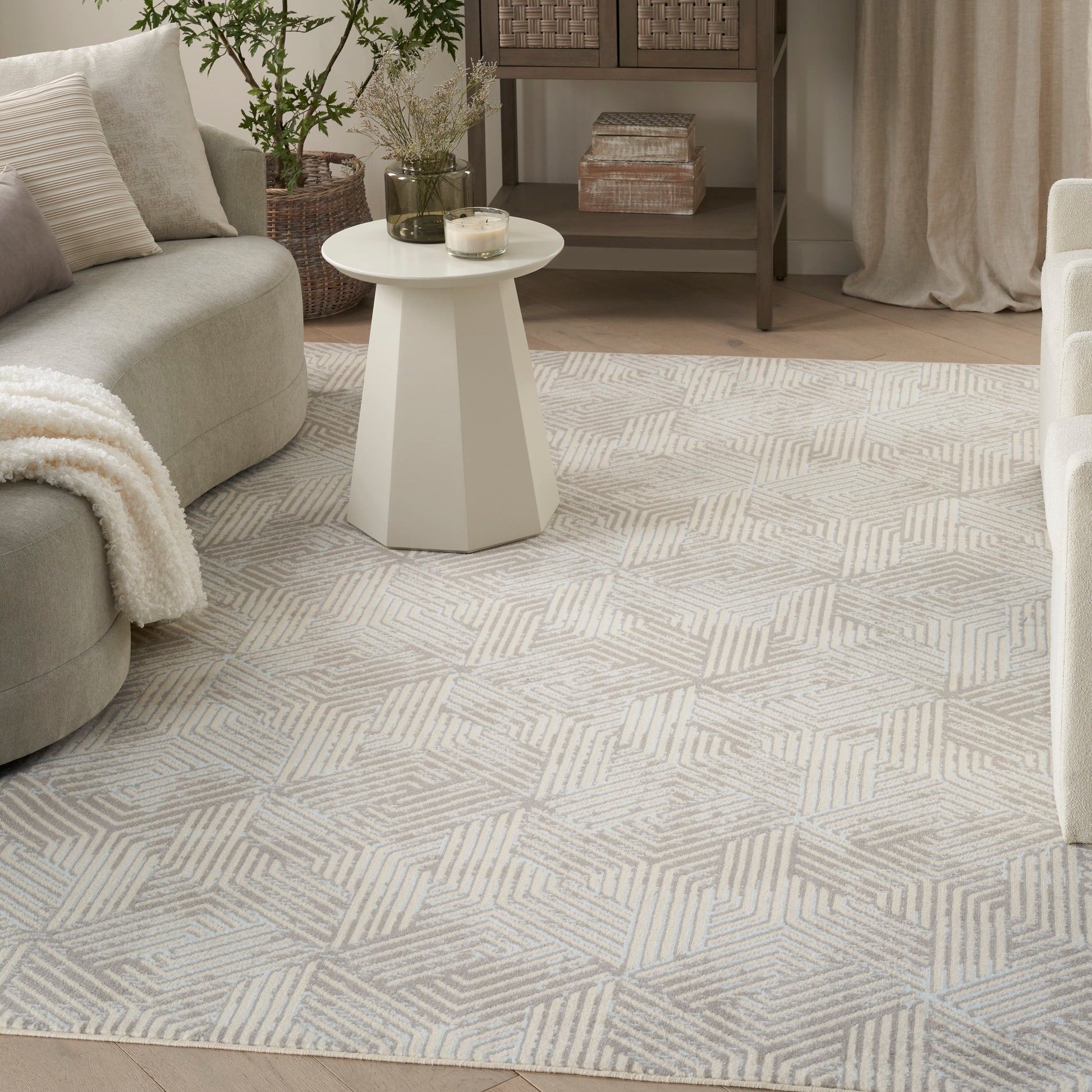 Light gray geometric patterned rug in a cozy living room with a white pedestal side table, sofa, throw blanket, and plants.
