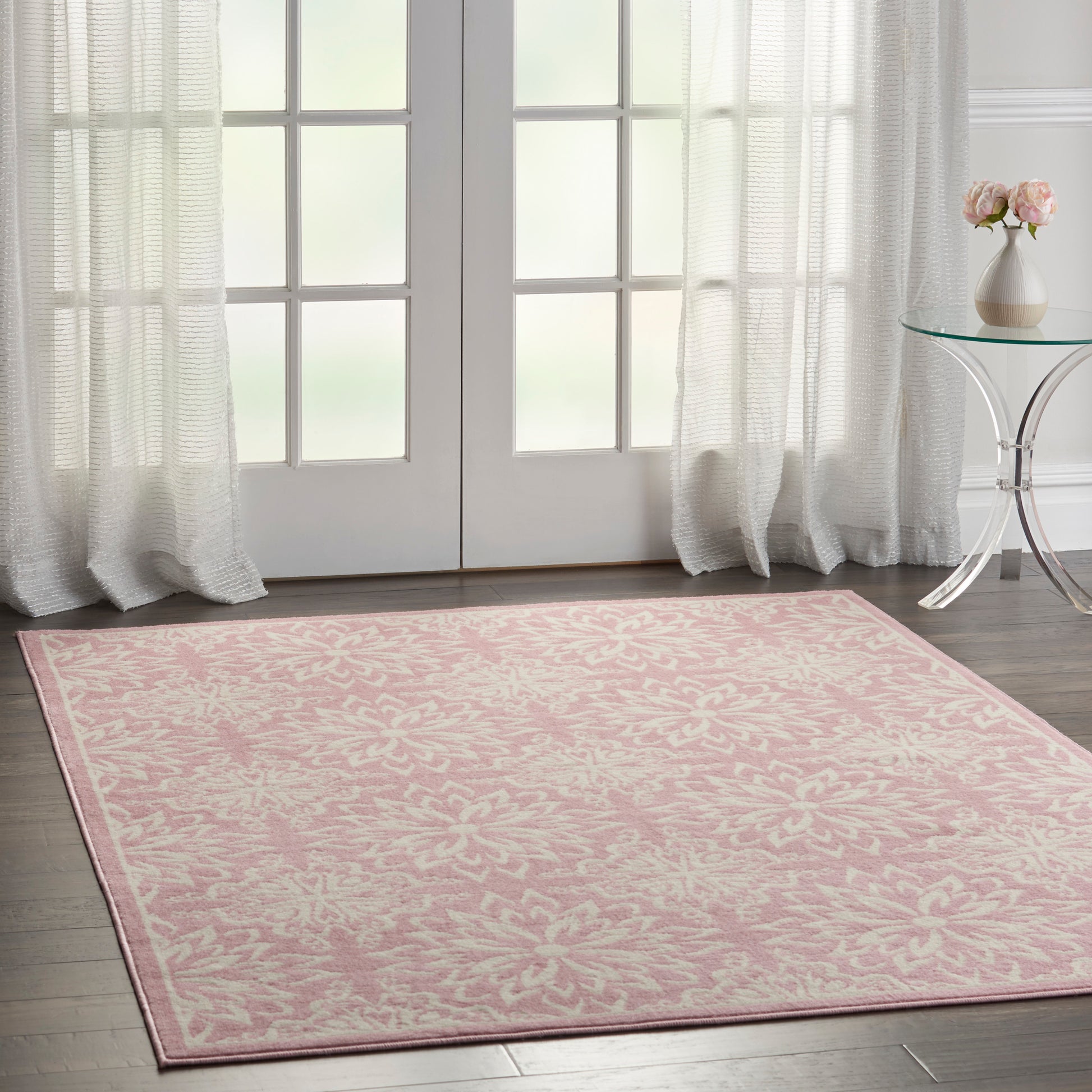 Rectangular pink area rug with white damask floral pattern, soft textile feel, placed on dark wood floor near glass doors.