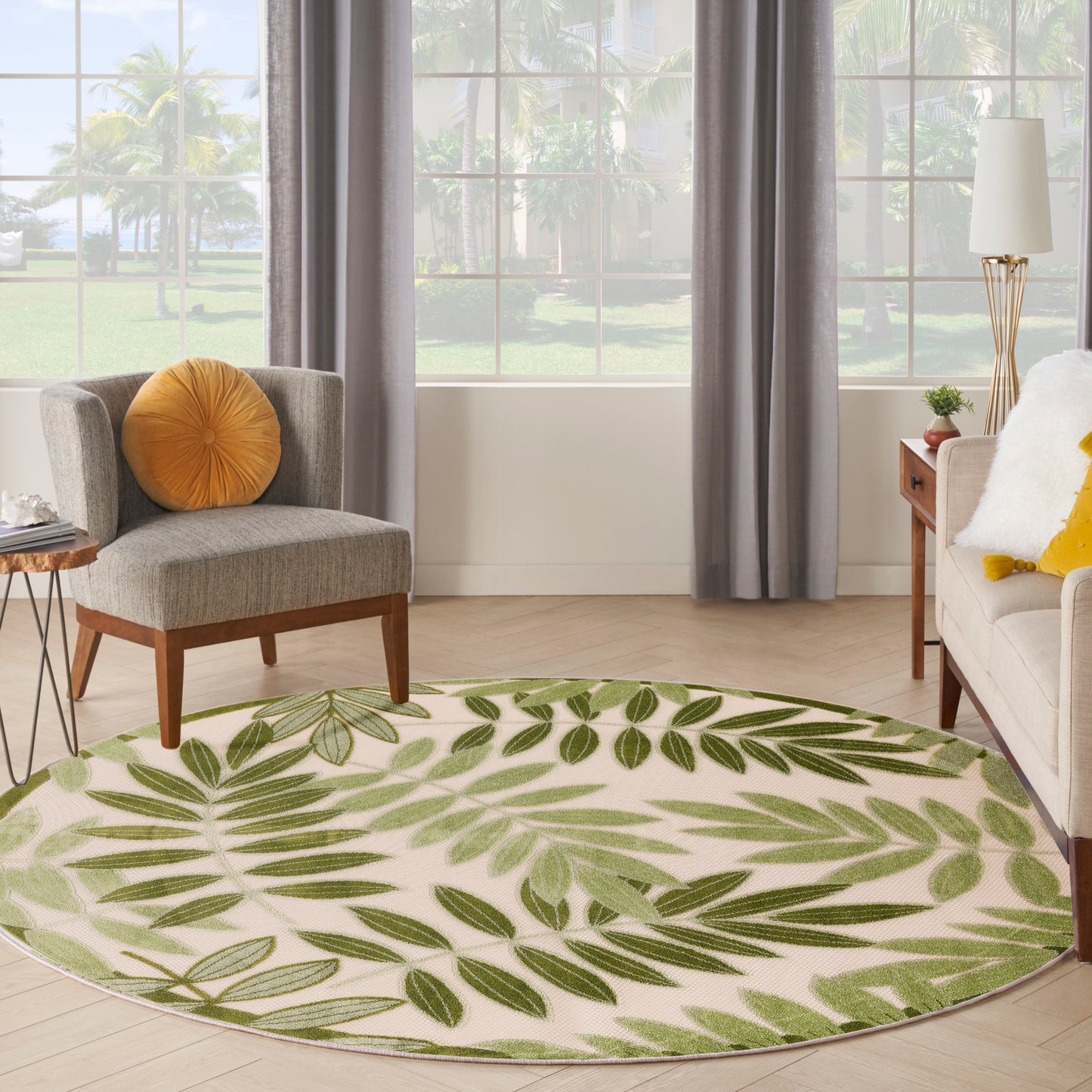 Round cream rug with green leafy pattern in a bright living room; gray armchair with orange cushion and beige sofa.