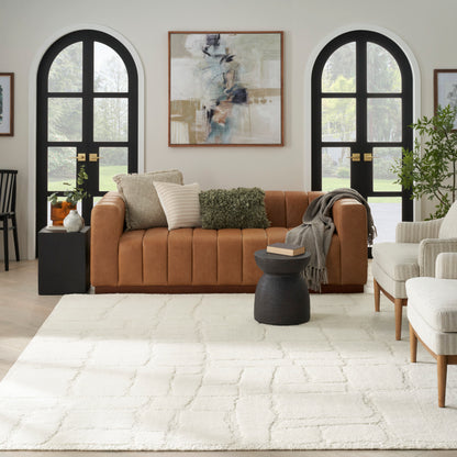 Warm tan leather sofa with beige and green cushions, cream shag rug, black arched windows, abstract wall art, gray throw; cozy living room.