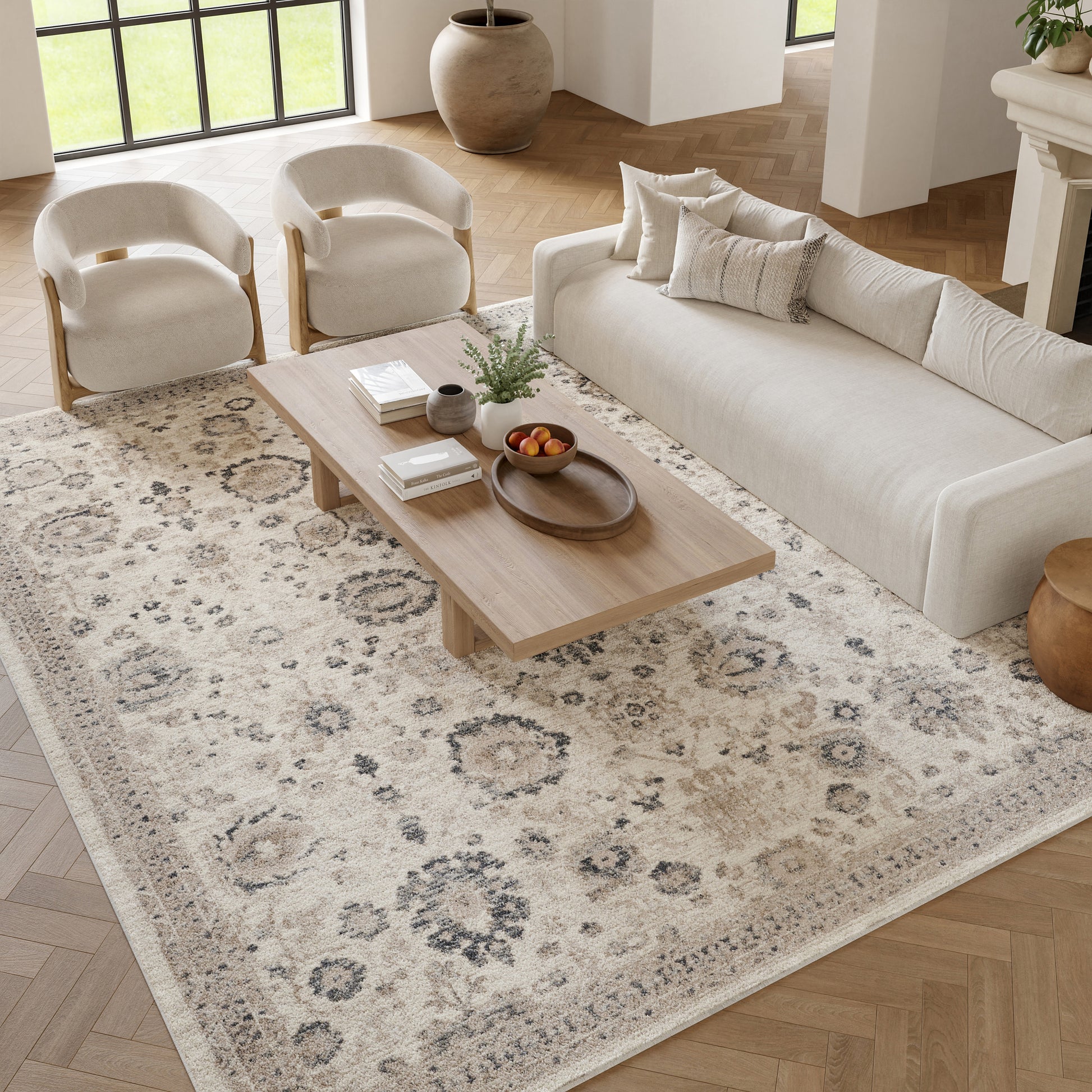 Neutral living room with beige fabric sofa and curved armchairs, light wood coffee table, ornate patterned area rug.