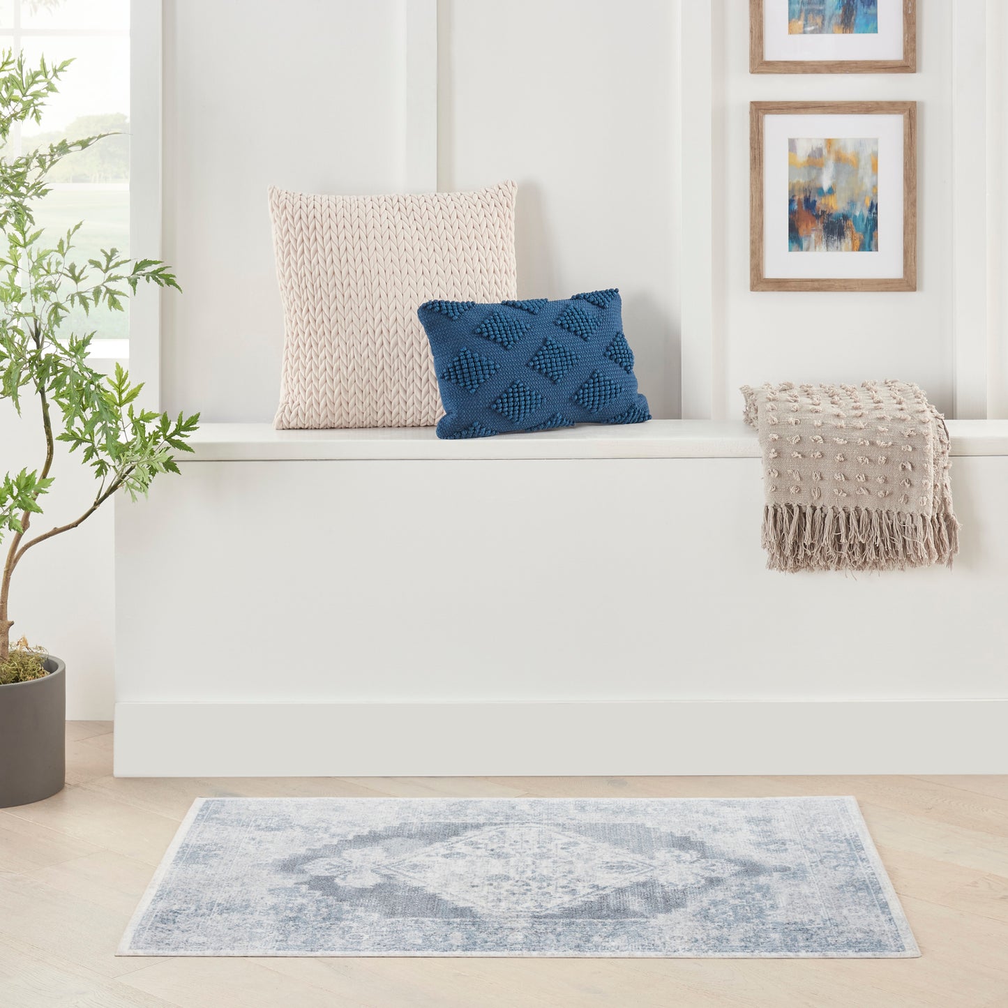 Beige knit cushion, navy textured geometric cushion, beige fringe throw on a white bench with a blue rug and framed art.