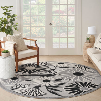 Round gray floral rug with black and white petals in a sunny living room with beige sofa, wooden chair, and French doors.
