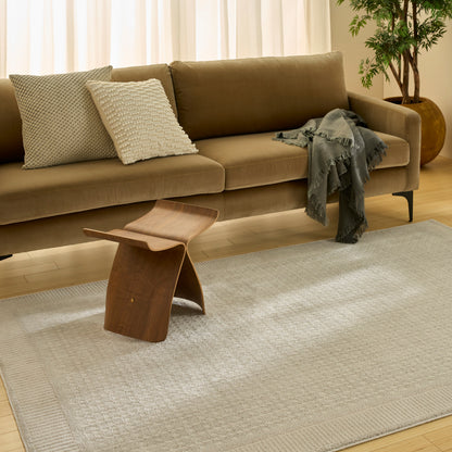 Light beige rug (4' x 6') in a warm living room setting, with a taupe sofa, gray throw, wooden curved stool and a potted plant.