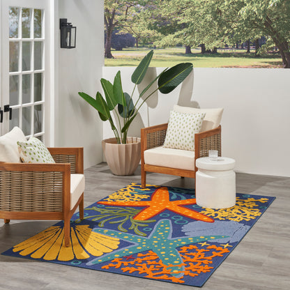 Outdoor blue rug with orange & yellow starfish pattern, shown in a setting with wicker chairs and large tropical plants.