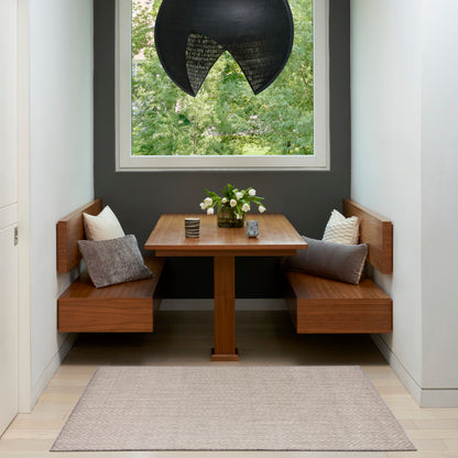 Wooden corner dining nook with pedestal table, built-in benches, gray walls, black pendant, beige rug, cushions, flowers.