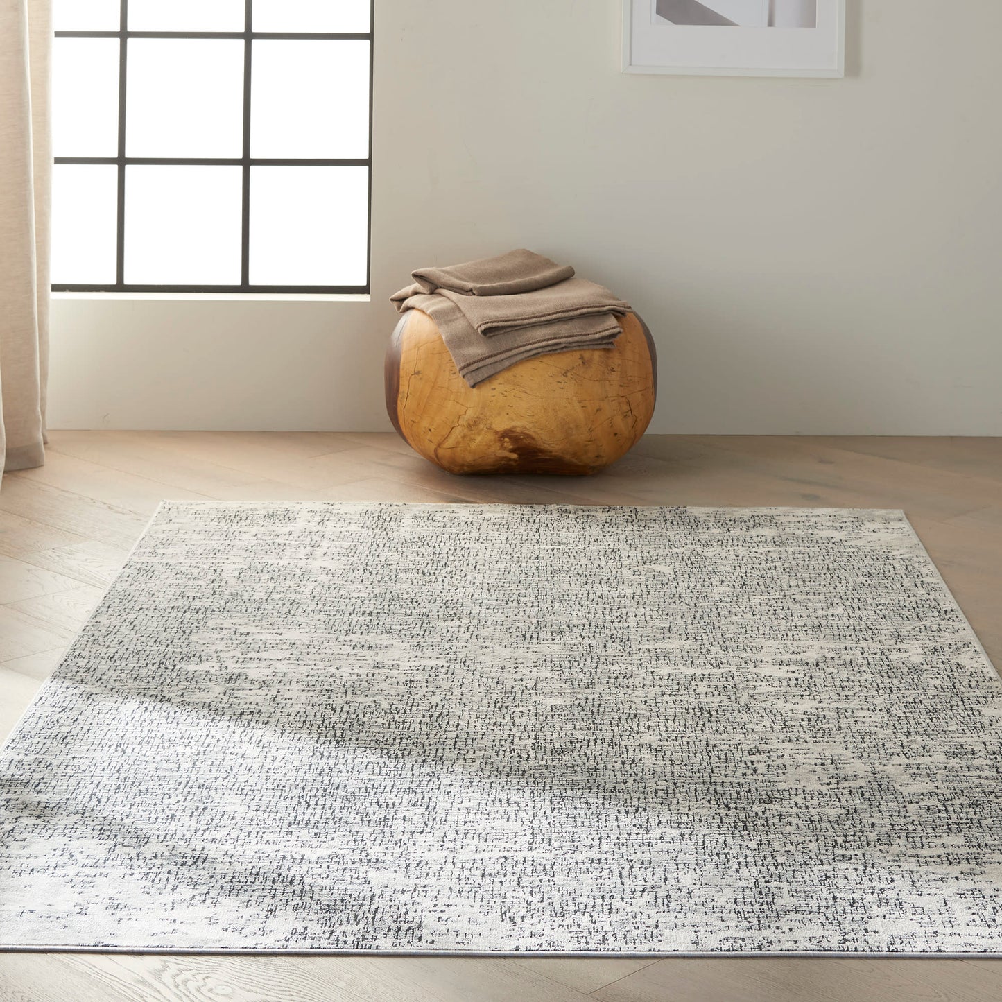 Light gray speckled area rug on hardwood floor with a round natural wood ottoman and beige blankets, sunlit by the window.