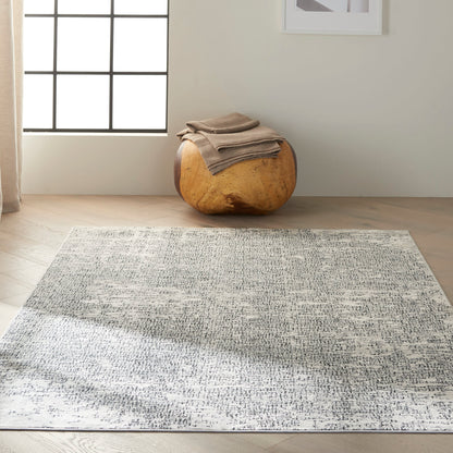 Light gray speckled area rug on hardwood floor with a round natural wood ottoman and beige blankets, sunlit by the window.