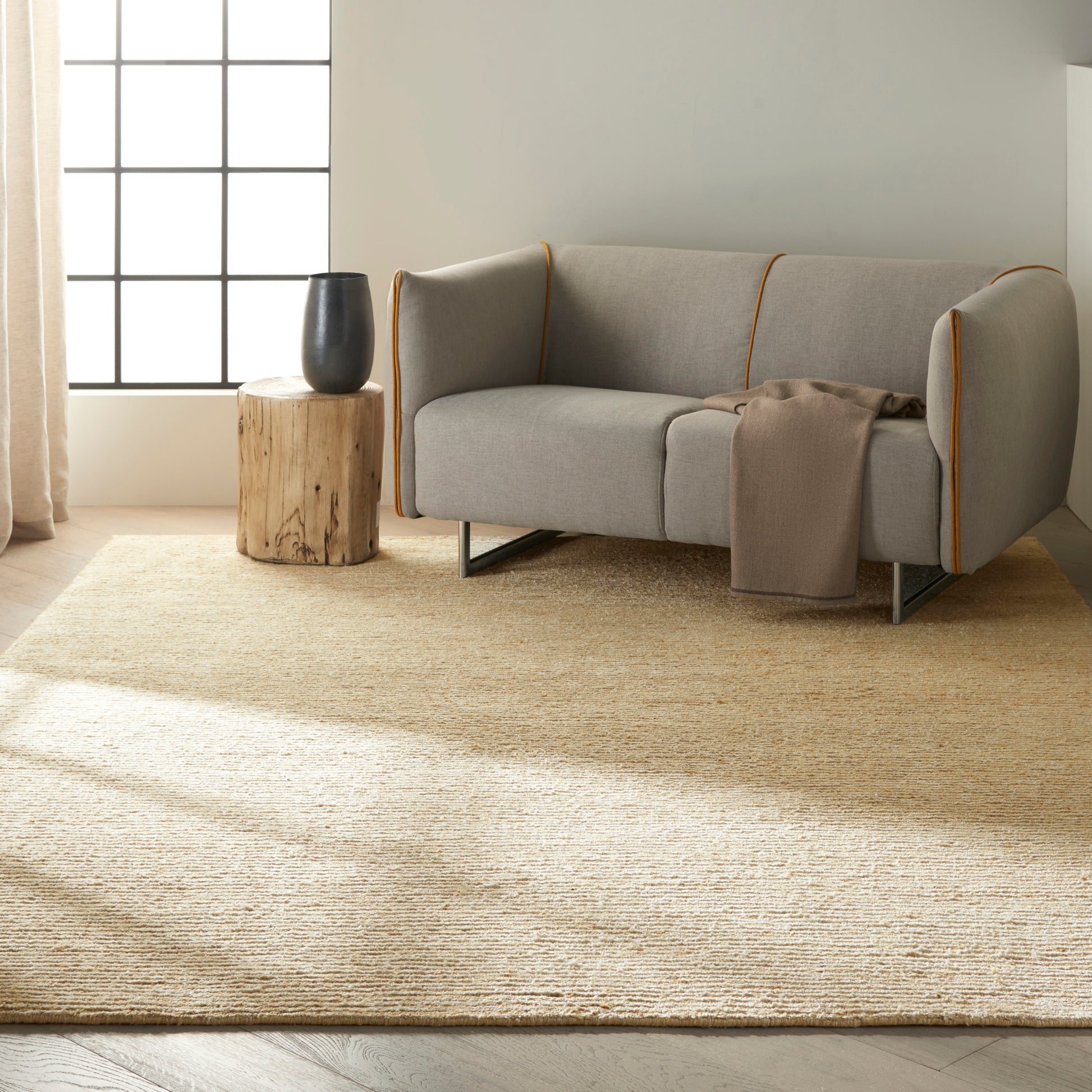 Light gray sofa with mustard piping, beige throw, sunlit living room with natural fiber rug and black vase on a stump table.