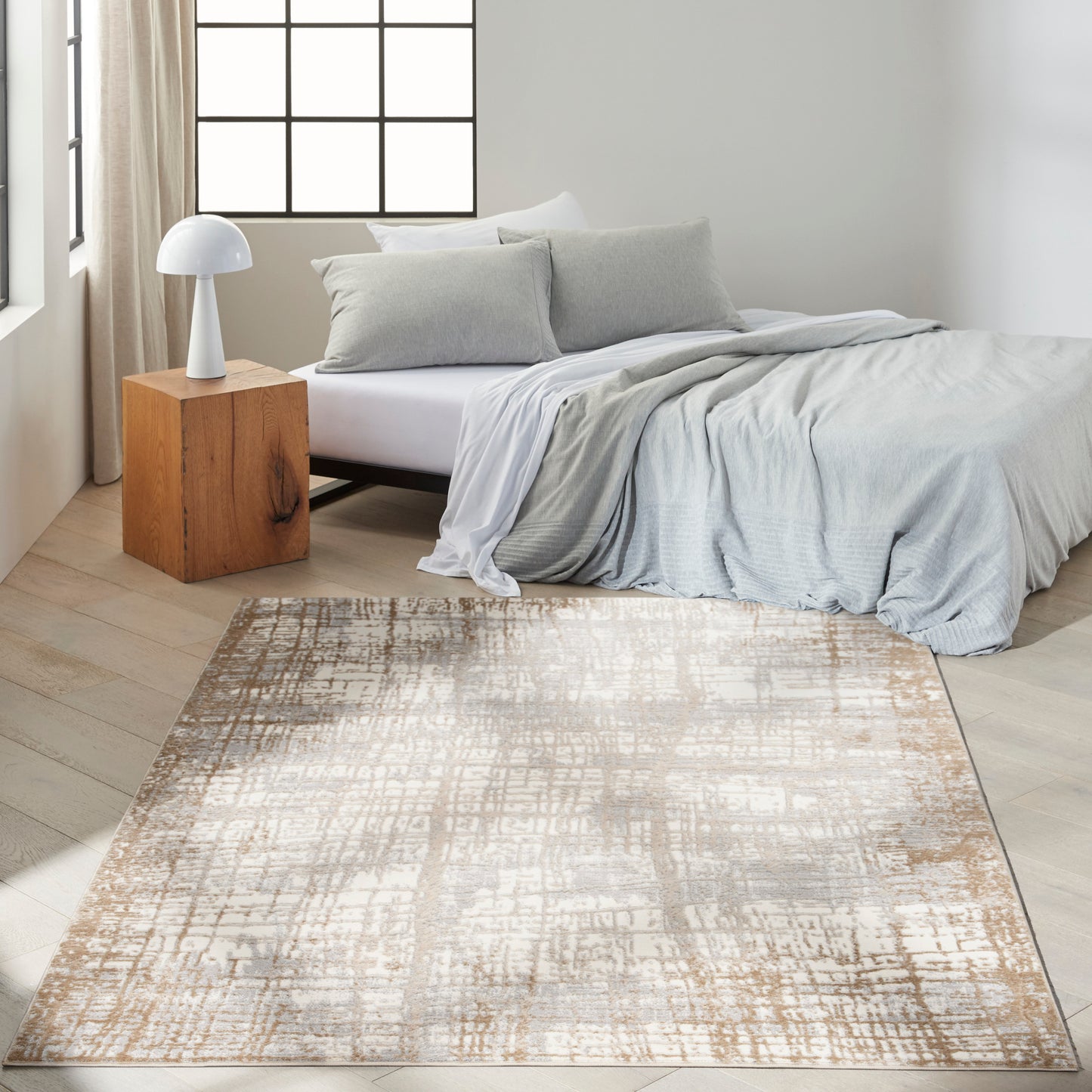 Minimalist bedroom with light gray bedding, white sheets, gray pillows, wooden cube nightstand, white lamp, and beige rug.