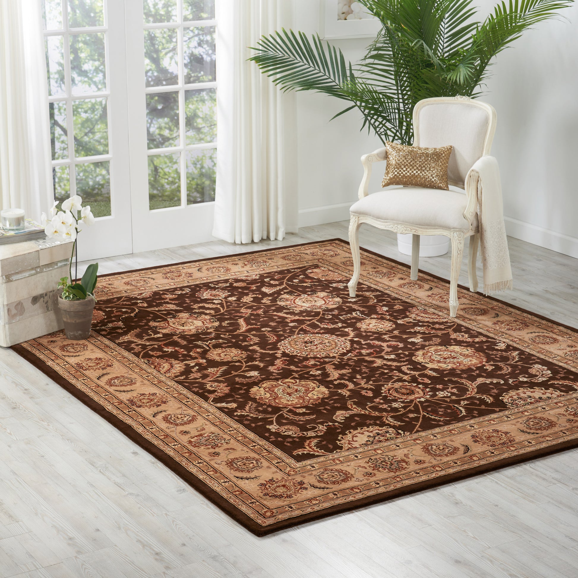 Oriental brown and beige rug with ornate floral border on light wood floor beside a cream upholstered chair.
