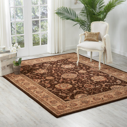 Oriental brown and beige rug with ornate floral border on light wood floor beside a cream upholstered chair.