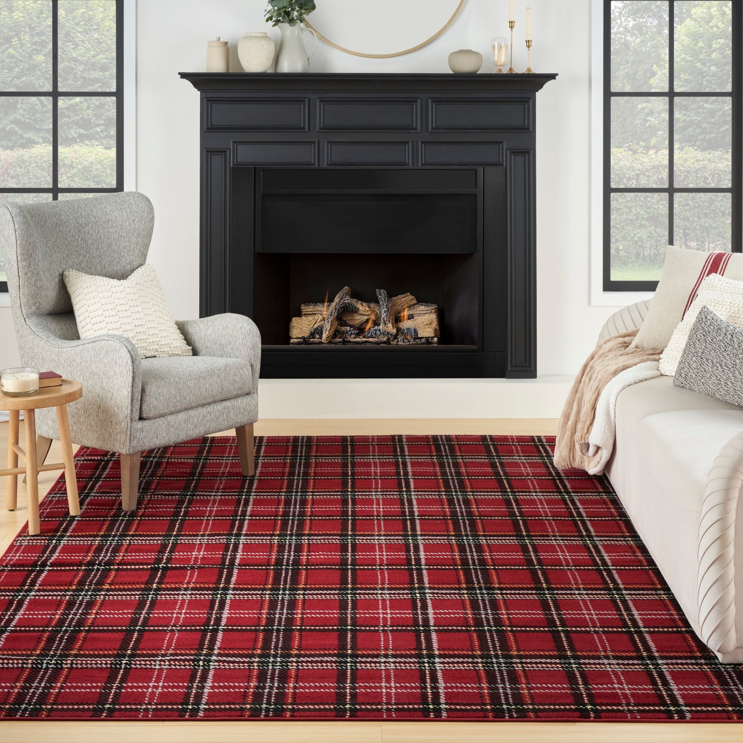 Cozy living room with a black wood fireplace, red plaid rug, gray upholstered chair, cream sofa, and round wall mirror.