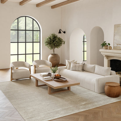 Neutral living room with beige fabric sofa, cream armchairs, natural wood coffee table, beige rug, arched window and fireplace.