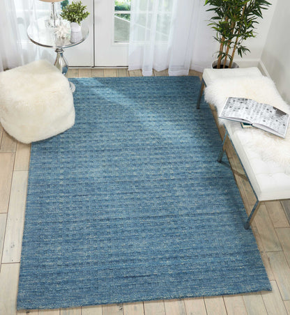 Blue textured woven area rug on light wood floor with white tufted bench, sheepskin throw, round glass table, and potted plants.