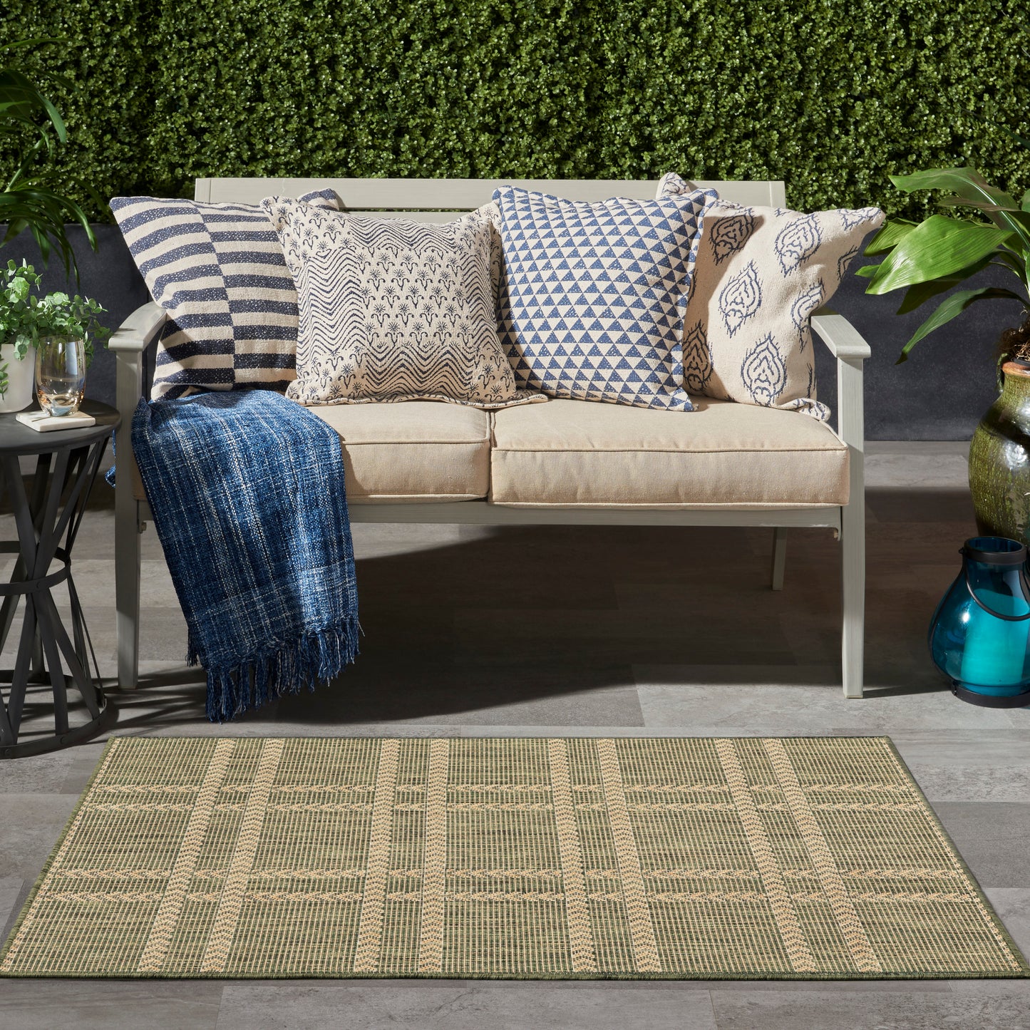Beige outdoor sofa with blue and white patterned cushions, a blue throw, on a woven beige rug amid greenery.