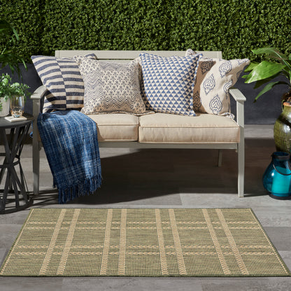 Beige outdoor sofa with blue and white patterned cushions, a blue throw, on a woven beige rug amid greenery.