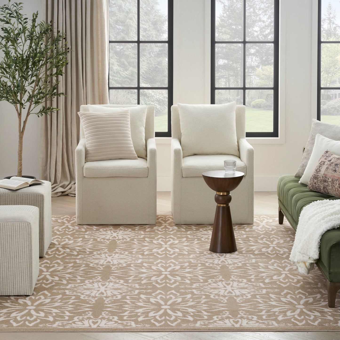 Two cream armchairs with cushions, round wooden side table, beige floral rug, green sofa, and a plant by large windows.