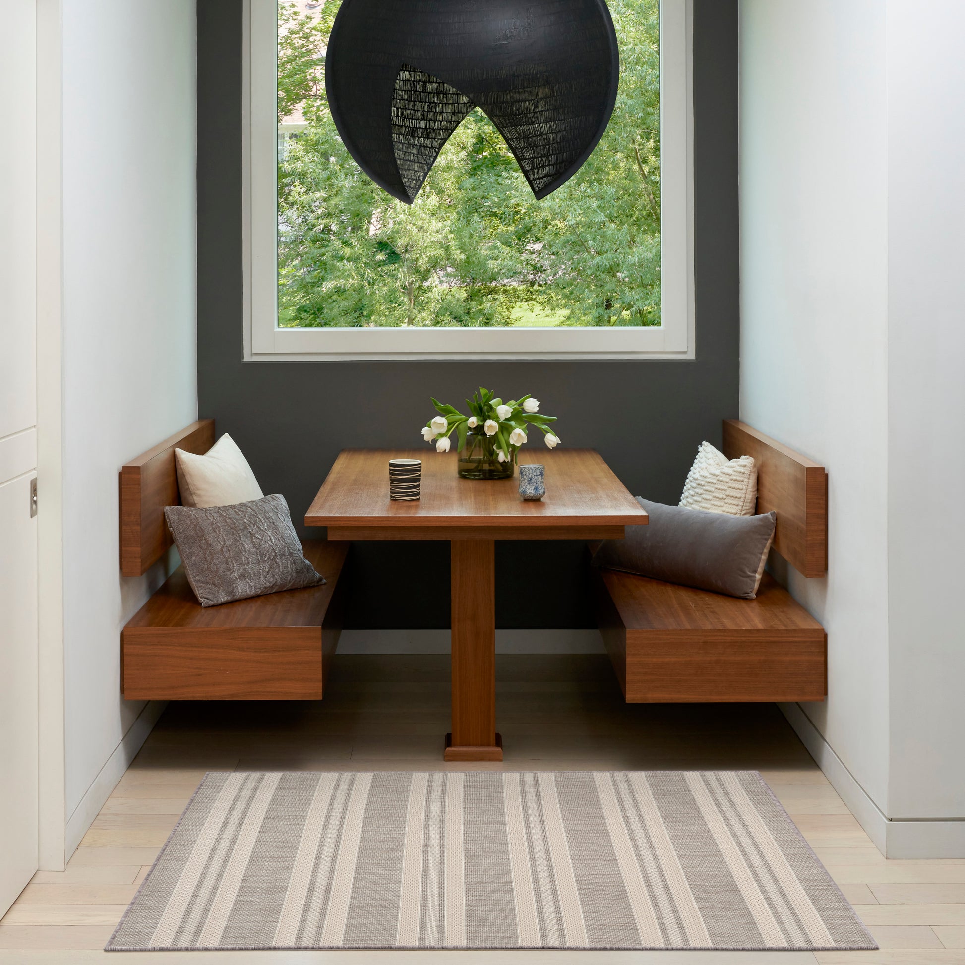 Cozy built-in wooden dining nook with rectangular table, gray walls, black pendant light, striped rug, and vase of flowers.