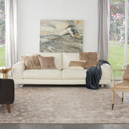 Beige patterned rug in a bright living room, with a cream sofa and textured tan pillows, a dark throw, and abstract gray-gold wall art.