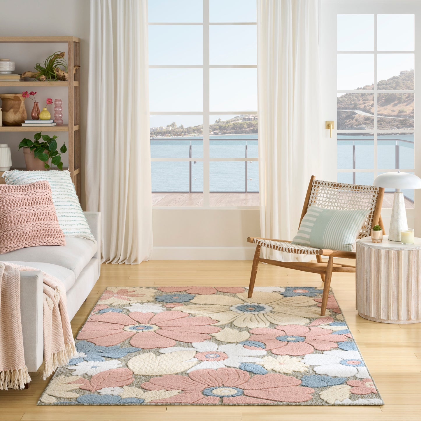 Coastal living room with pastel floral rug, light wood furniture, cream sofa, wicker chair, round side table, sea view.